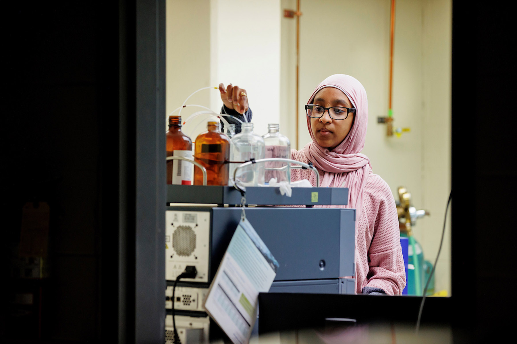 Scientist in hijab and glasses works with laboratory equipment, examining bottles near scientific machinery.